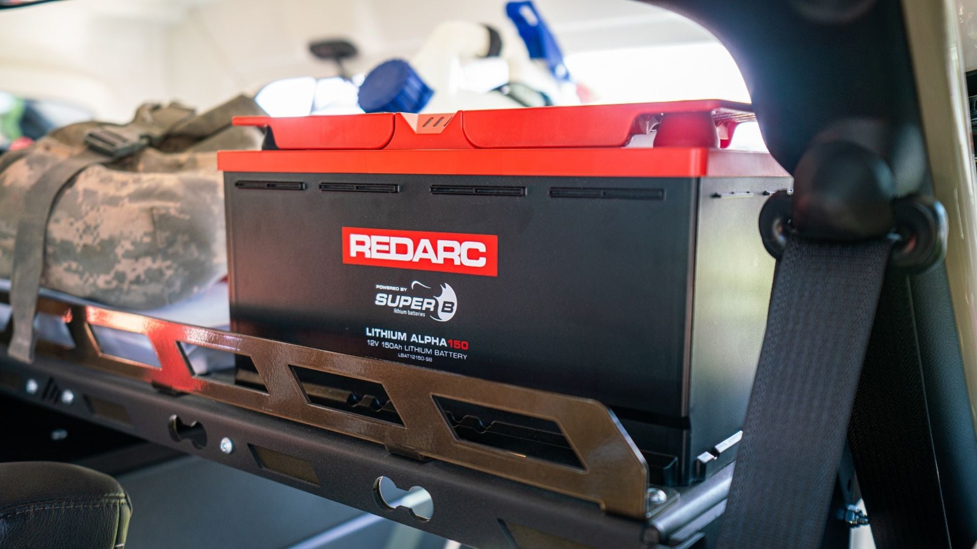 Close-up of a REDARC Lithium Alpha 150 battery secured inside a vehicle, with camping gear visible in the background