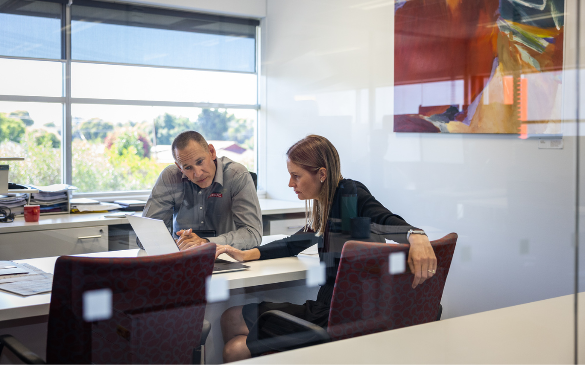 A man and woman sit at a desk in an office. They're looking at a laptop and talking.
