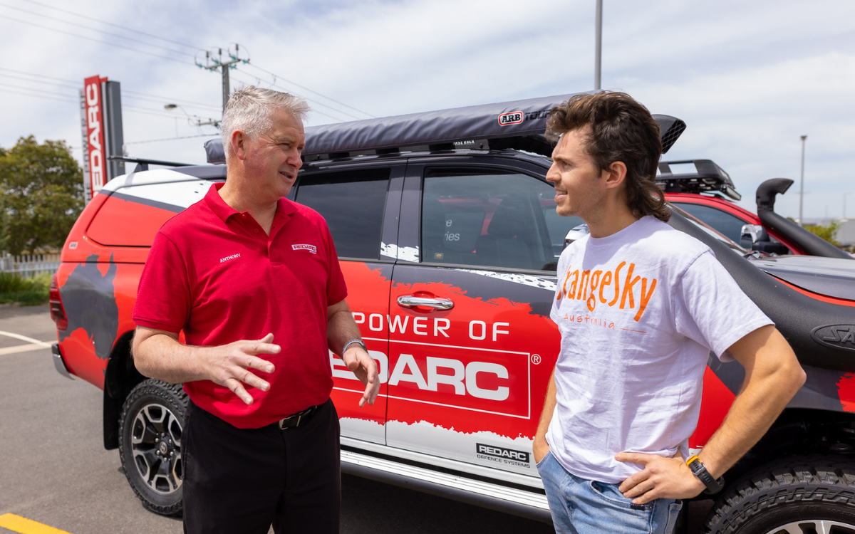 Two men smiling and talking in front of a REDARC branded 4WD. One man wears a red shirt and the other a white shirt that says 'Orange Sky'.