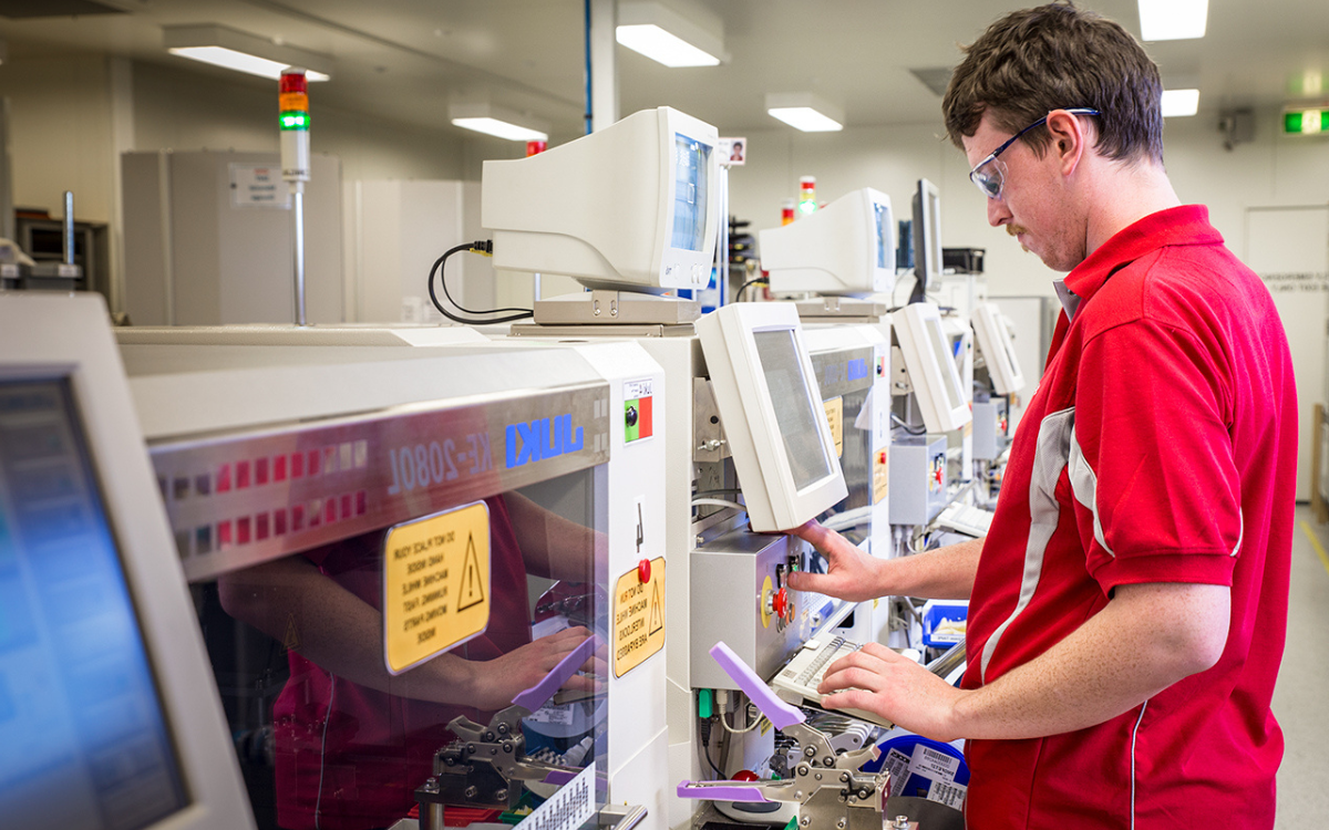 A man in a red shirt pressing a button on a screen that is attached to a line of surface mount technology machines.