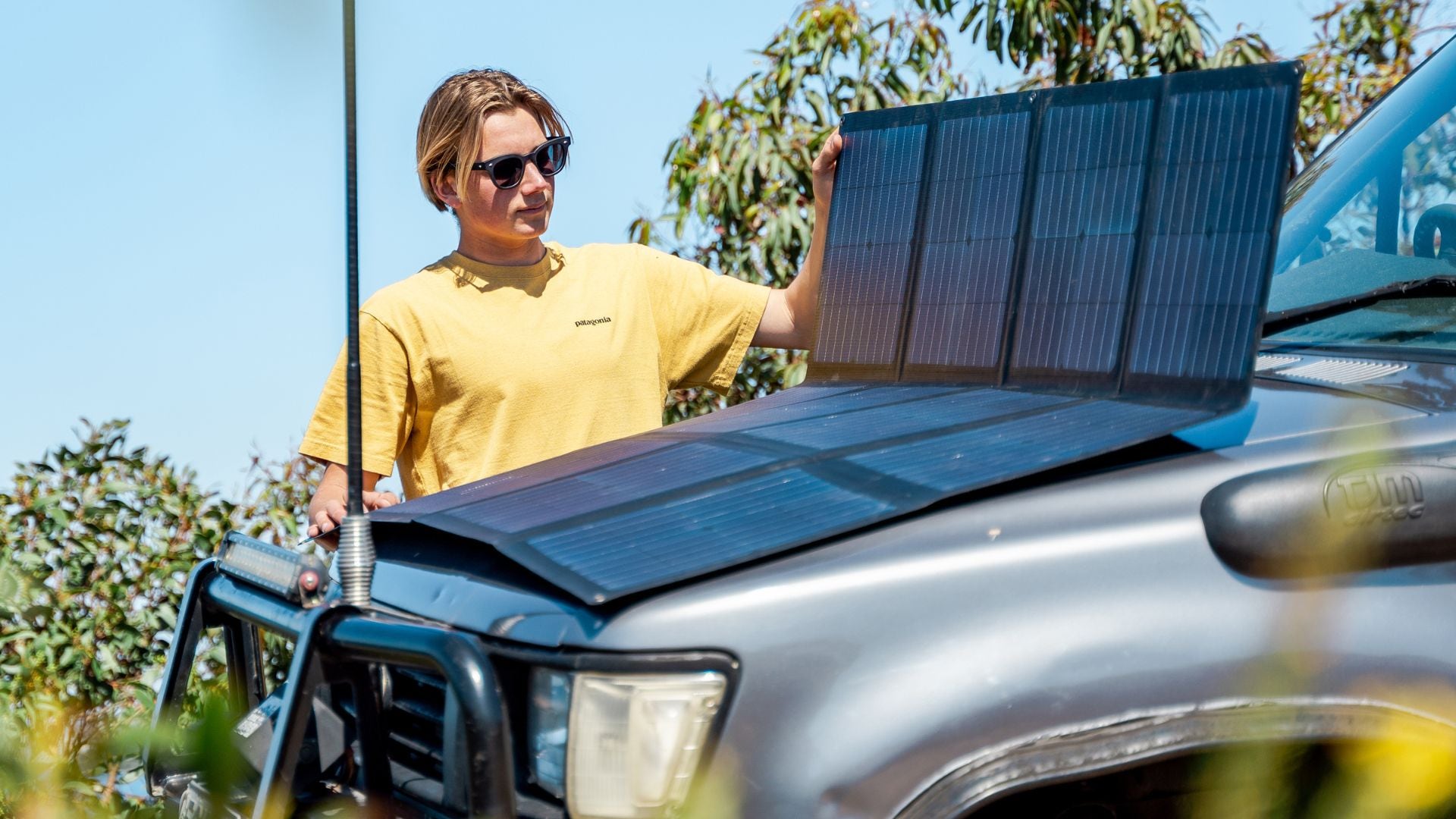 Woman setting up portable solar panels near an off-road vehicle by a lake on a bright, sunny day Woman setting up portable solar panels next to an off-road vehicle by a grassy lakeside on a sunny day